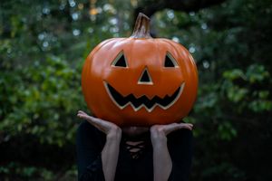 Someone wearing black holds up a carved Halloween pumpkin with classic triangular eyes and zig-zag smile against greenery.