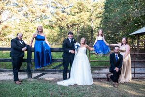 A wedding party in formal attire poses together by a wooden fence at an outdoor rustic venue.