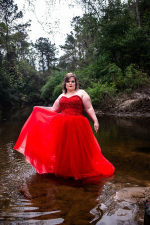 A stunning red ballgown dress flowing in a woodland creek setting with pine trees and natural scenery in the background.