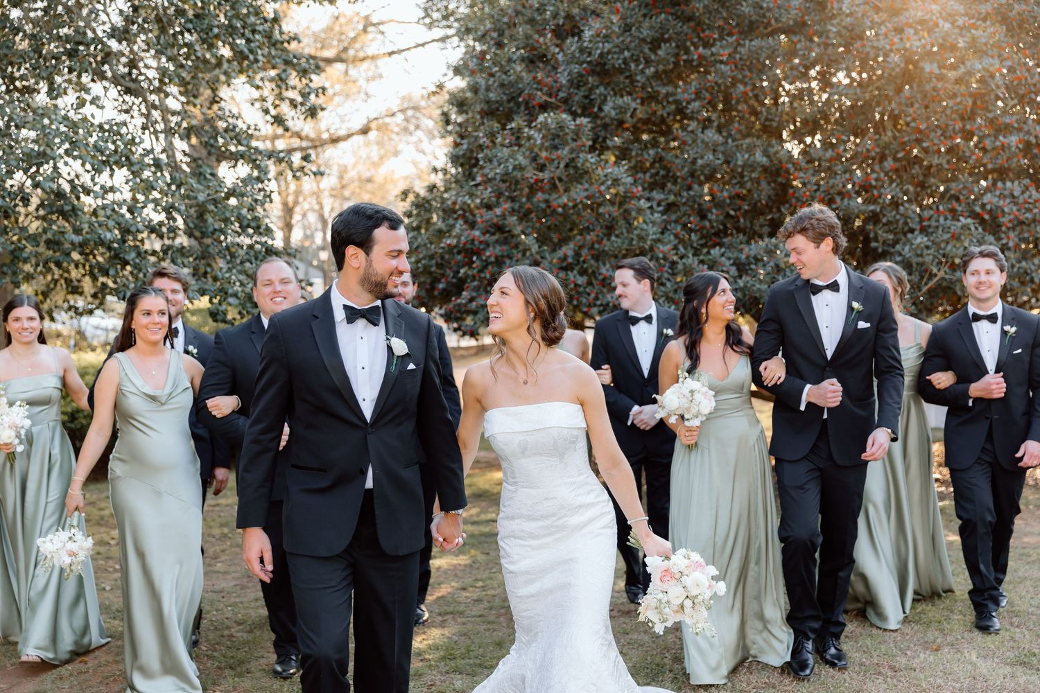 Bride and groom walk with wedding party in sage bridesmaid dresses and black tuxedos on a sunny outdoor lawn at Naylor Hall.