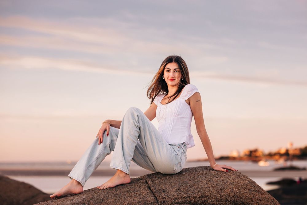 Person in white dress poses on rocky beach at sunset while ocean waves crash in background.