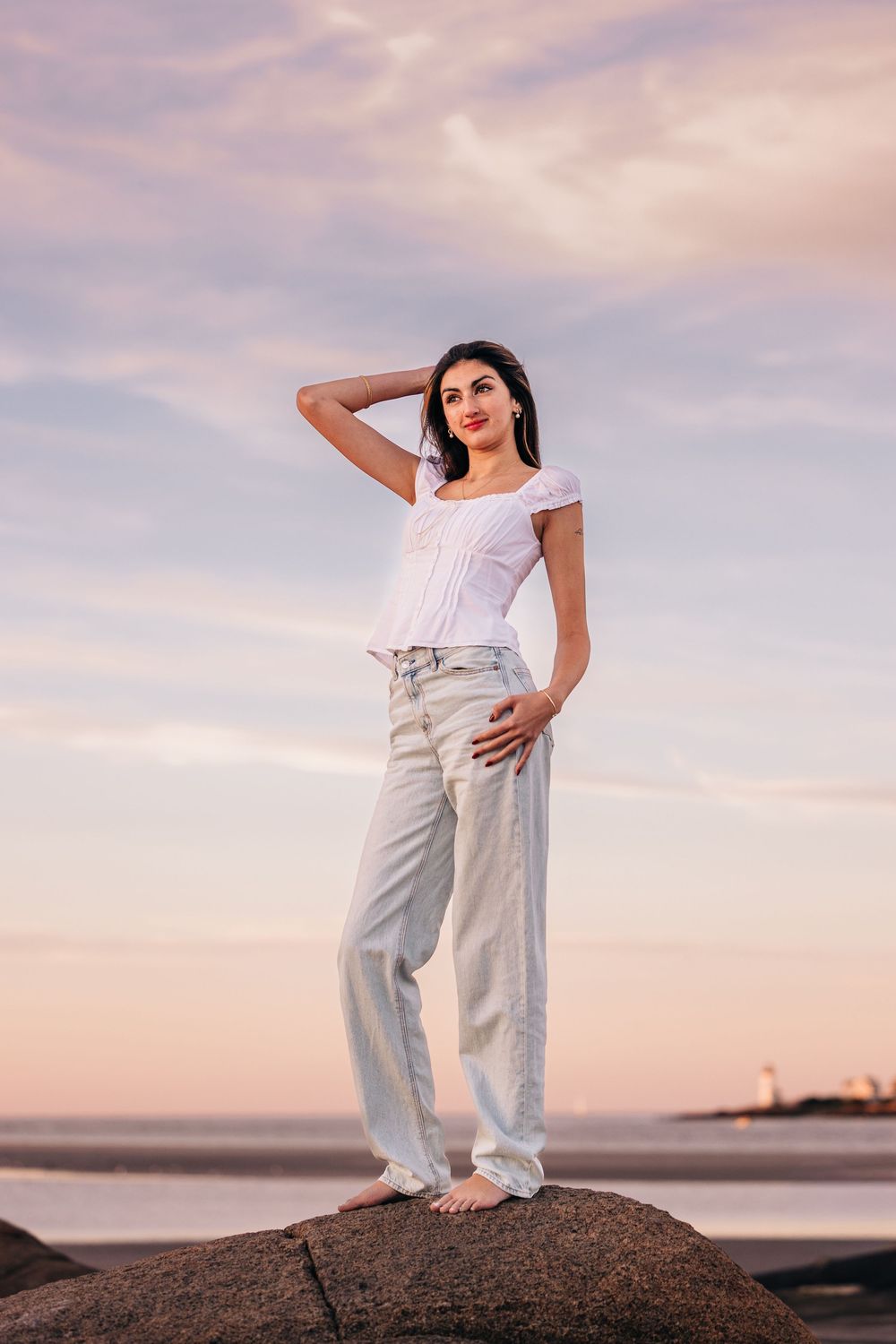 Person in white top and light jeans poses on coastal rock formation during sunset with pink cloudy sky.