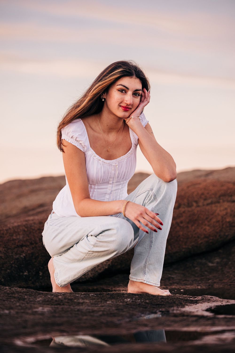 A person in a white dress sits gracefully on rocky terrain at sunset, creating a serene coastal portrait series.