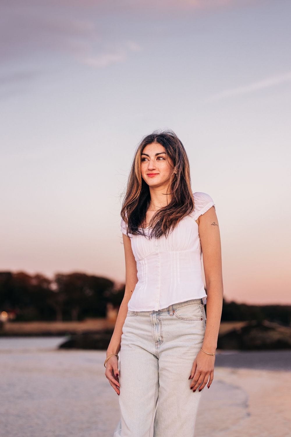 A woman in a white top and light jeans poses at sunset by the water's edge.