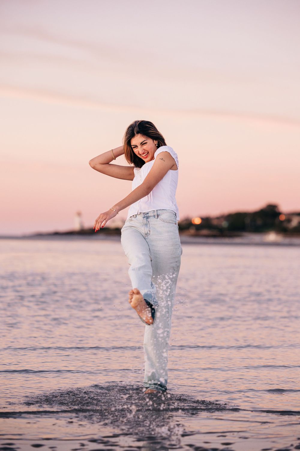 A dancer in white performs graceful leaps and splashes in shallow ocean water at sunset.