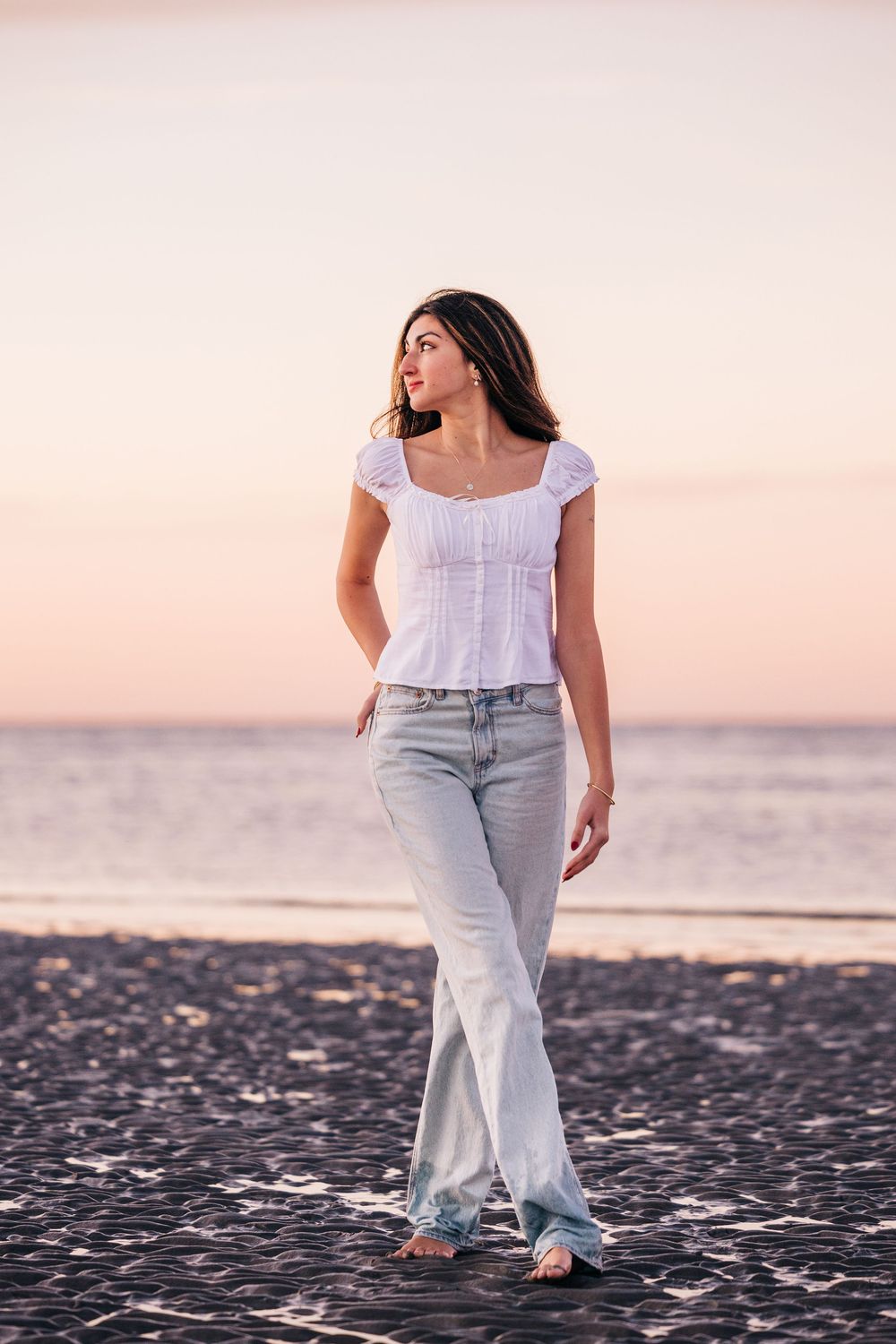 A person wearing a white top and light jeans walks along a dark beach at sunset with ocean waves in the background.