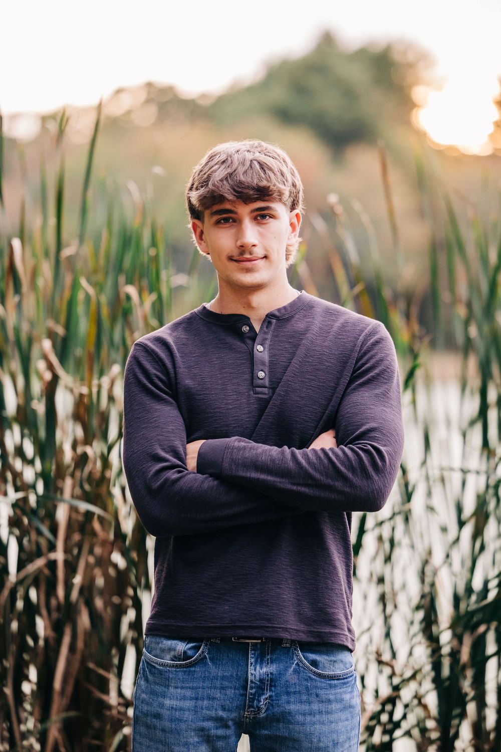 Portrait of someone in a navy henley shirt and jeans standing with crossed arms in front of tall reeds at sunset.