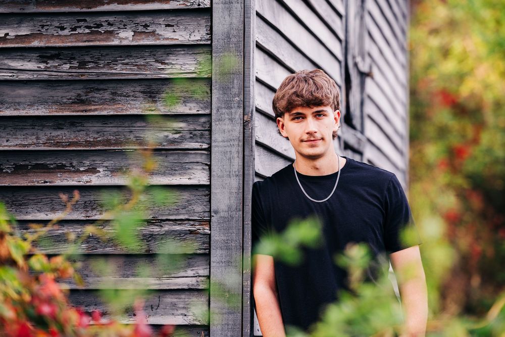 A person in a black t-shirt poses against a rustic wooden wall with greenery and autumn colors in the background.