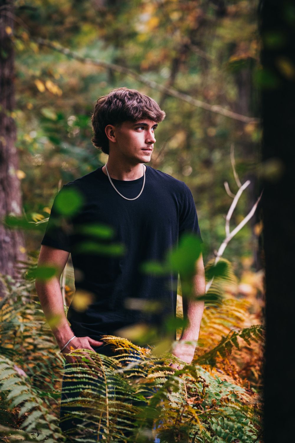 A figure in a black shirt poses among autumn ferns in a moody outdoor portrait setting.