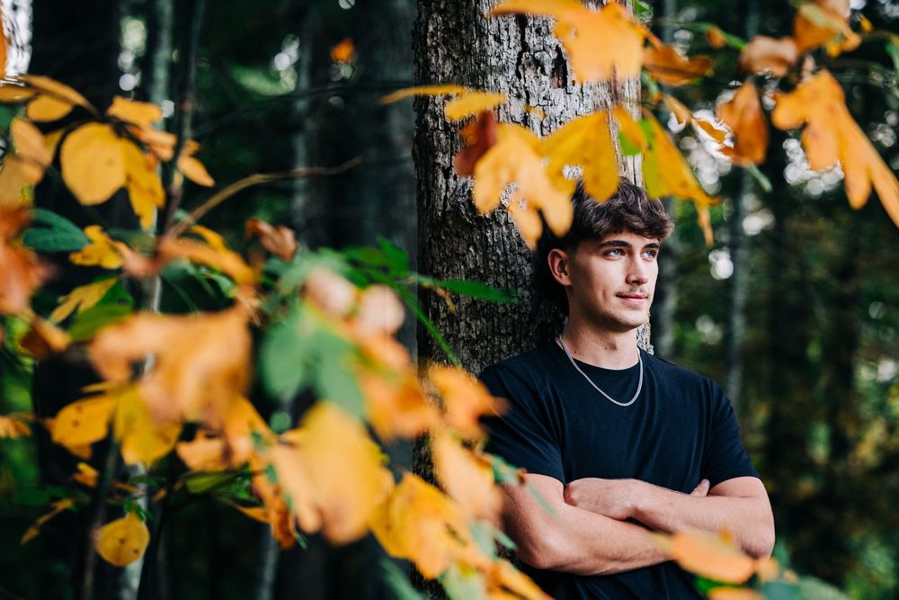 Person in black shirt stands in autumn forest with golden leaves and natural bokeh lighting effect in background.