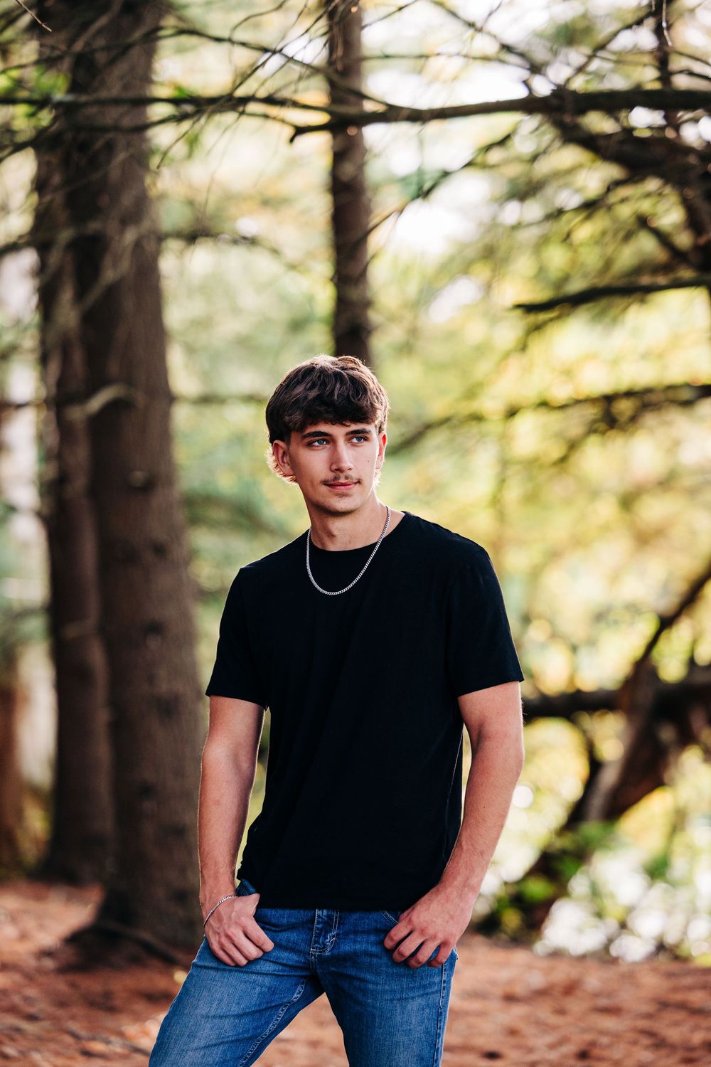 Portrait series in forest setting with natural lighting and pine trees in background, wearing black t-shirt and jeans.