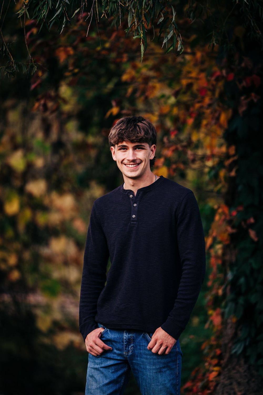 Someone wearing a black sweater and jeans stands against a backdrop of fall foliage in natural lighting.