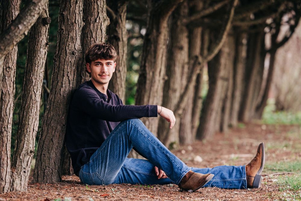 A person in casual attire and a hat sits on fallen leaves against tree trunks in a wooded area.