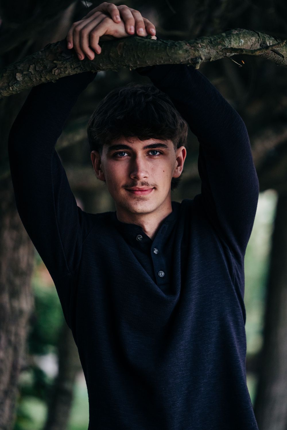A casual portrait under tree branches features dark clothing against a shadowy natural background.