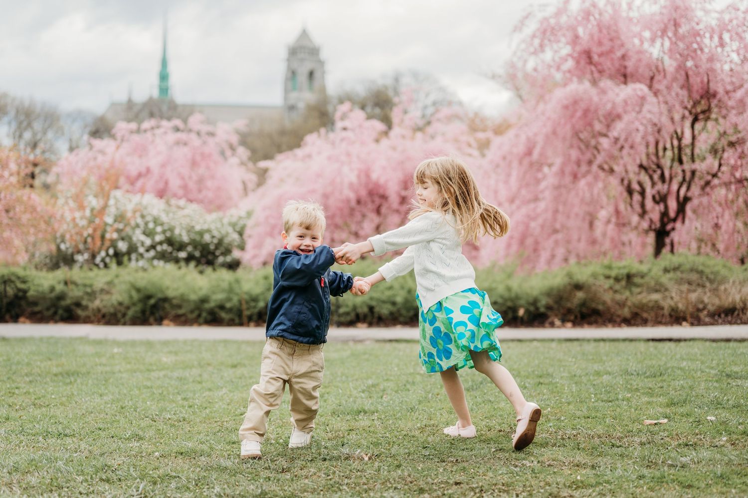 Two children dance and play together on a grassy lawn with beautiful pink cherry blossom trees and a church spire in the background.
