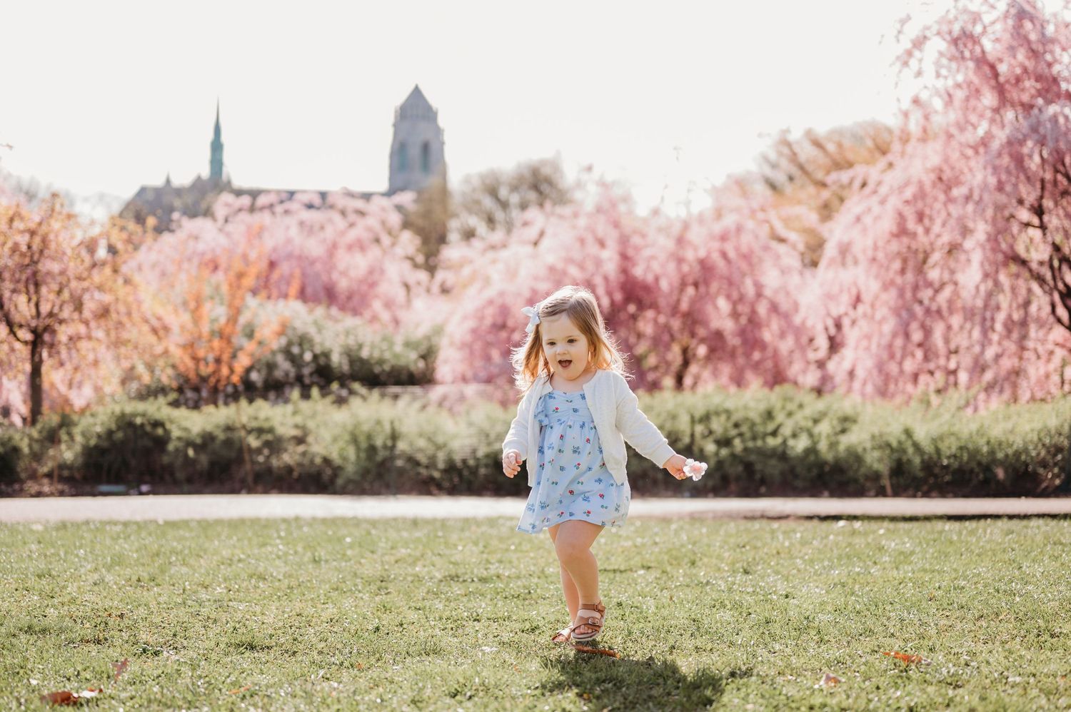 Small child in dress running on grass in front of cherry blossom trees and cathedral spires in background at Branch Brook Park in Newark.