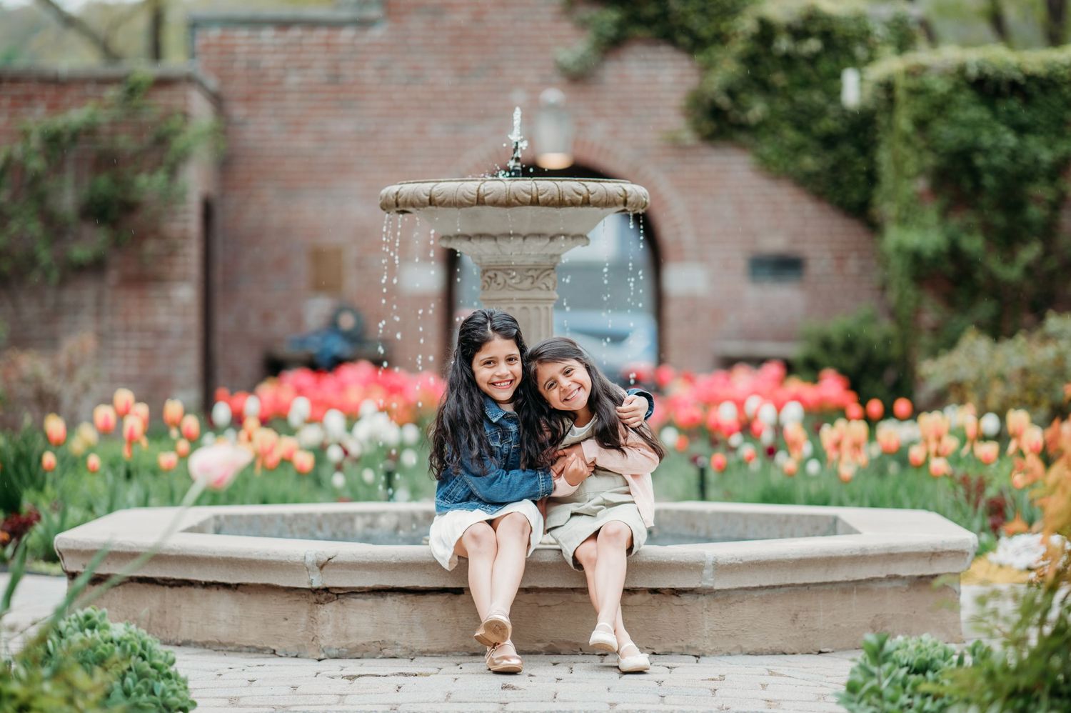 Two sisters sitting together at a stone fountain in a garden surrounded by blooming tulips in Montclair.