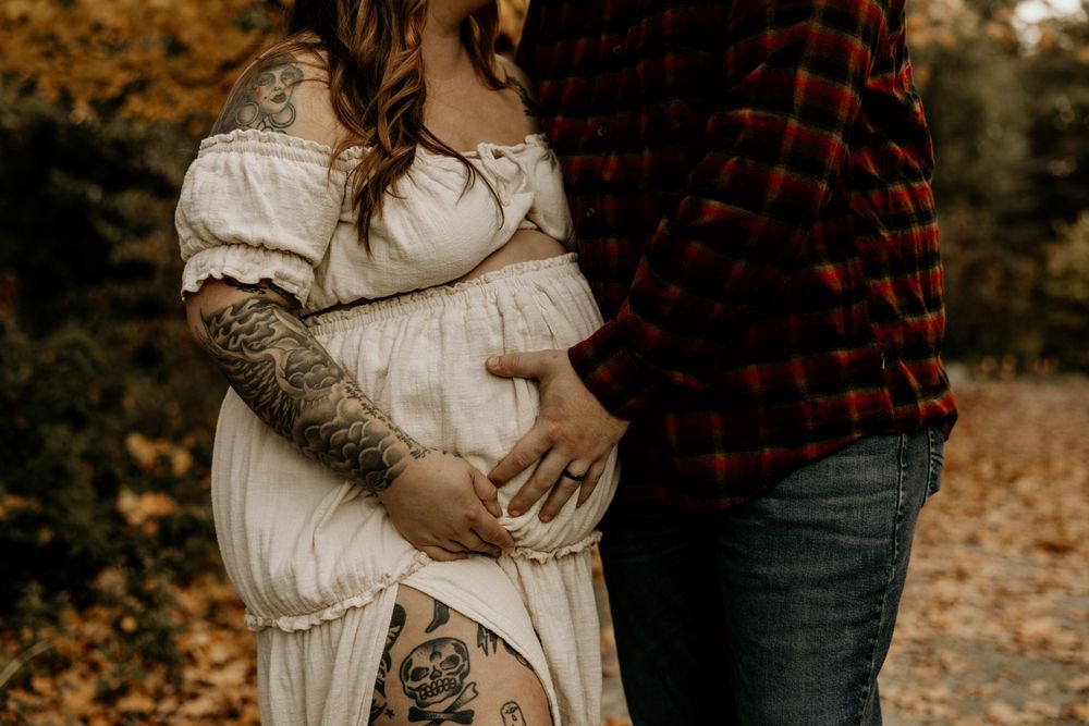 A couple in autumn embrace while wearing a white dress and plaid shirt, with visible arm tattoos on display.