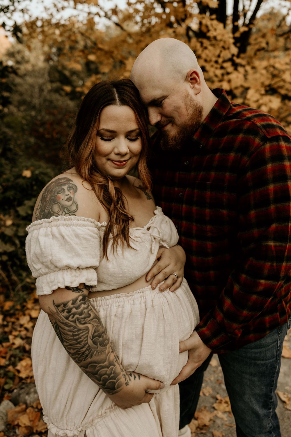 Intimate autumn maternity photo showing a couple in a white dress and red plaid shirt embracing amid fall foliage.