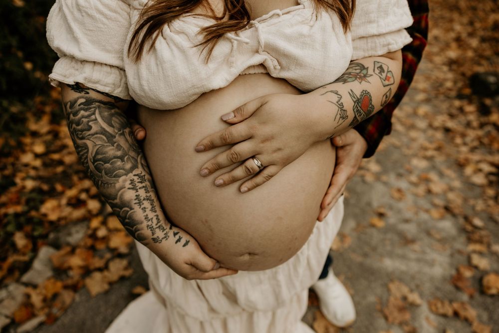 Close-up shot of tattooed hands cradling a pregnant belly in a white dress against autumn leaves on the ground.