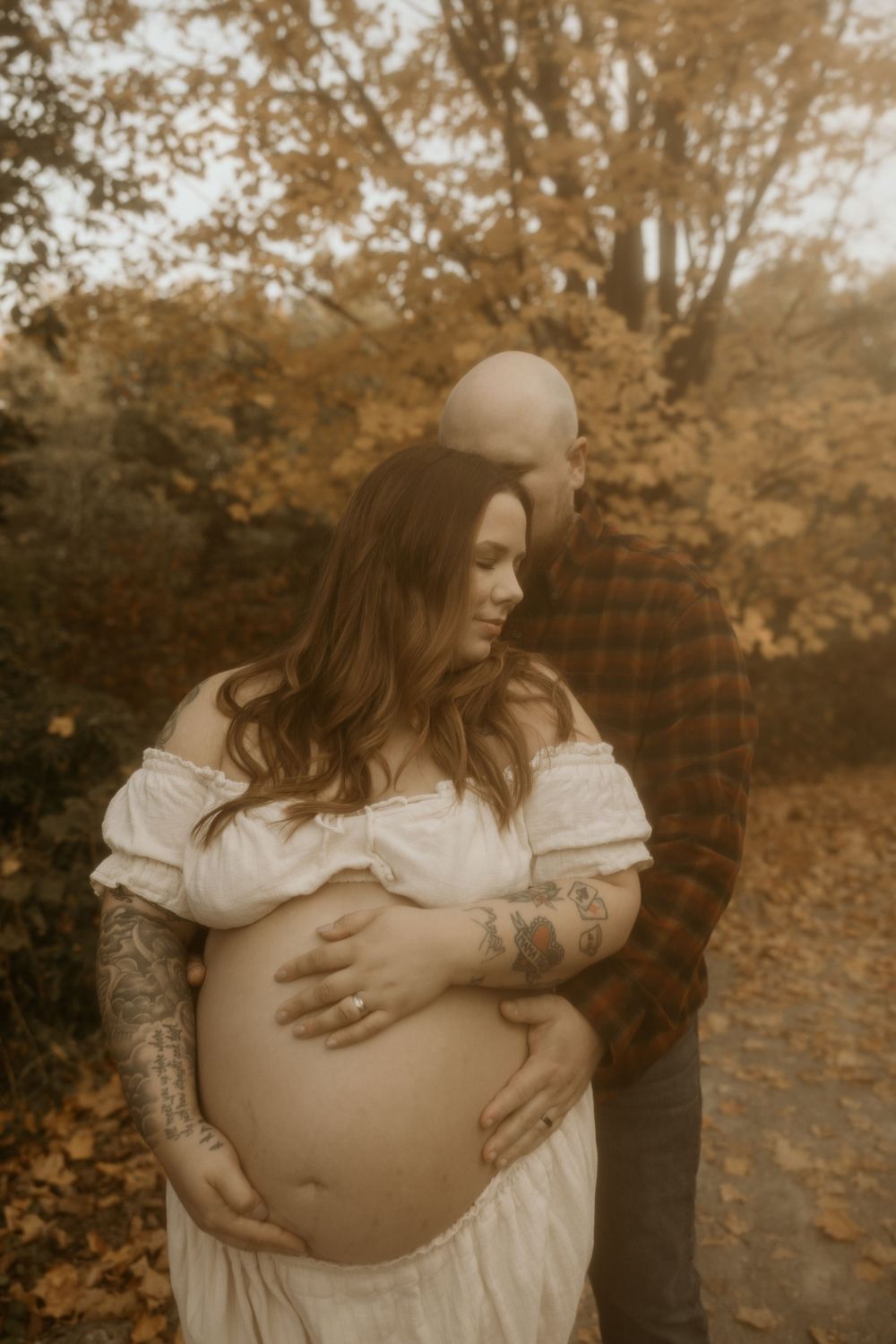 Sepia-toned maternity photo outdoors showing a pregnant woman in a white dress with arm tattoos during autumn.