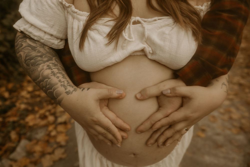 A heart shape formed by hands over a pregnant belly, with autumn leaves visible in the background.