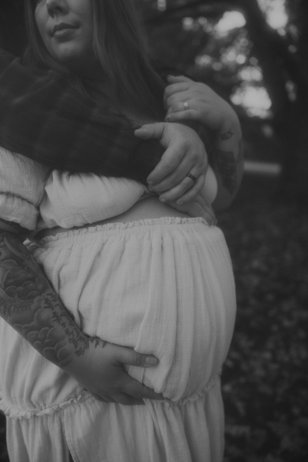 Black and white photo of a pregnant woman in a flowing white dress holding her belly outdoors in soft natural light.