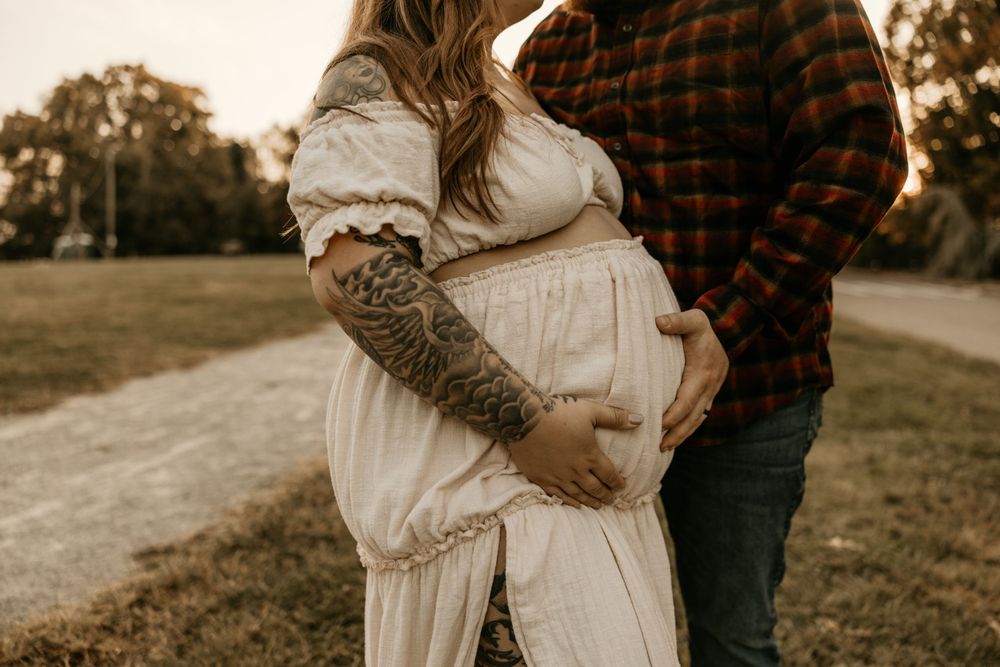 A couple embraces during a maternity photo session at sunset, with the person wearing flannel showing a tattooed arm.