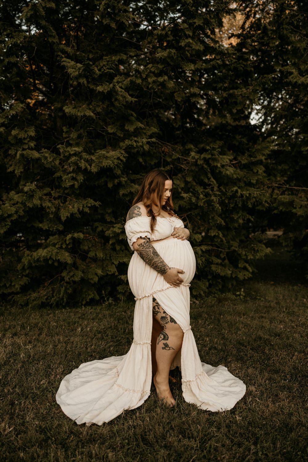 A pregnant woman in a flowing white dress stands outdoors in a natural setting with green foliage in the background.
