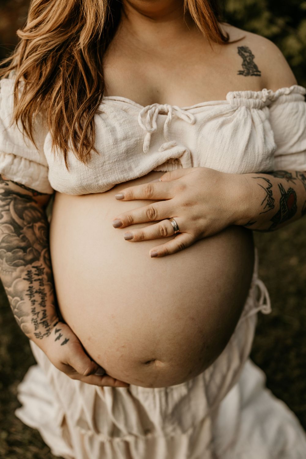 Tattooed expectant mother in white dress cradling baby bump in sunlit maternity photo.