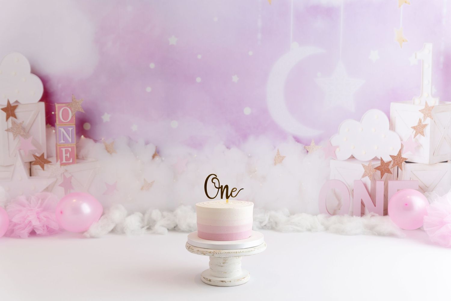 Small white cake with gold 'One' topper displayed on pedestal against dreamy pink and white cloud-like backdrop with stars.