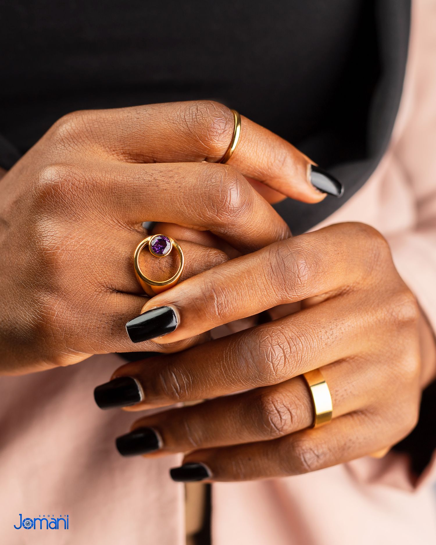 Close-up of hands with black nails wearing gold rings including a purple gemstone ring.