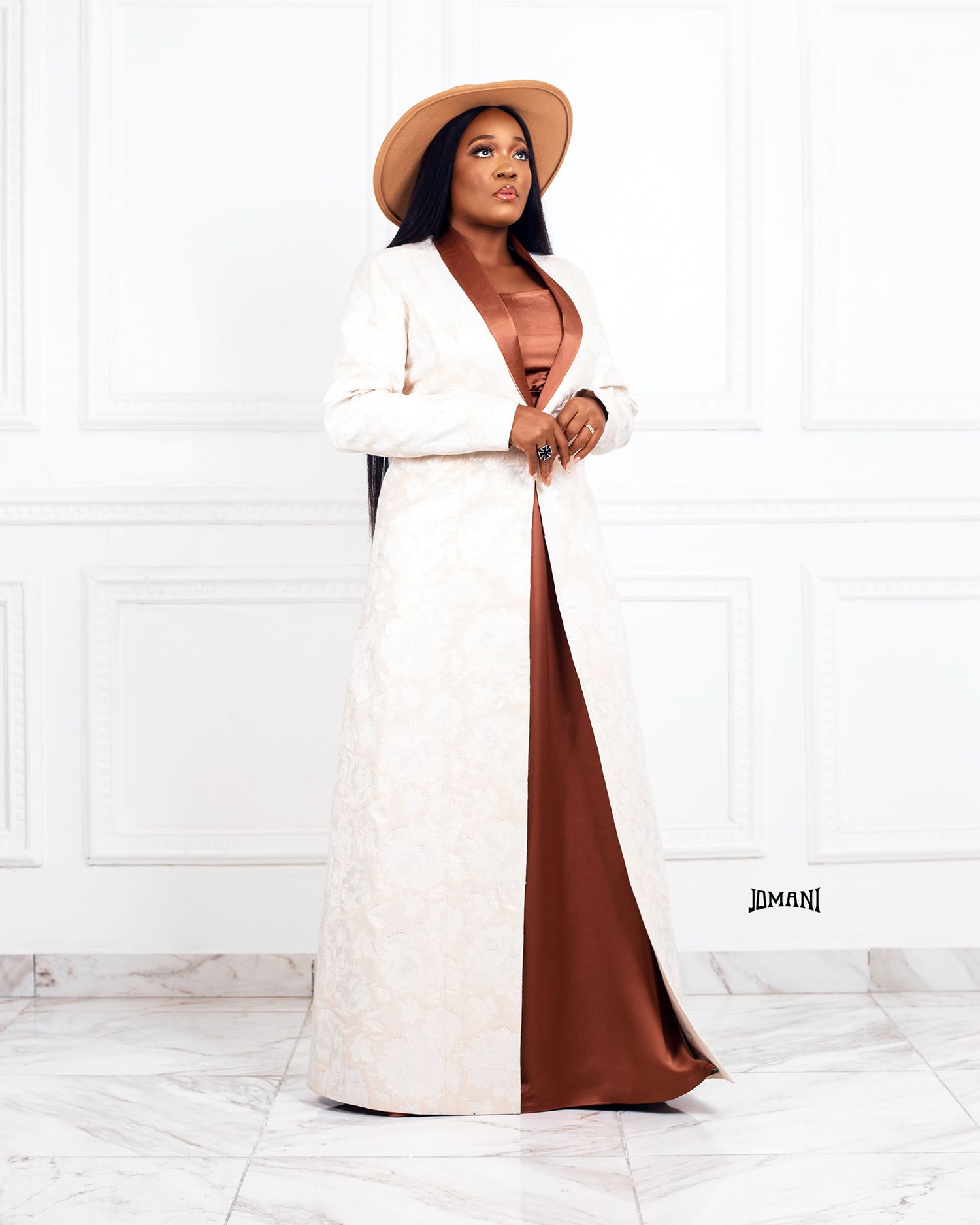 Woman in white lace coat and brown satin maxi skirt wearing a tan wide-brim hat poses elegantly.
