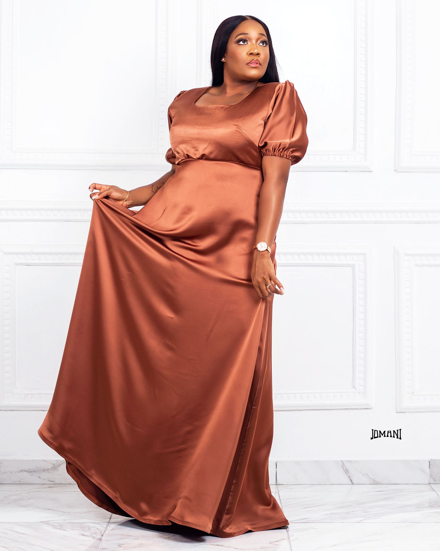 Woman in a flowing brown satin maxi dress with puff sleeves poses gracefully in a bright studio.