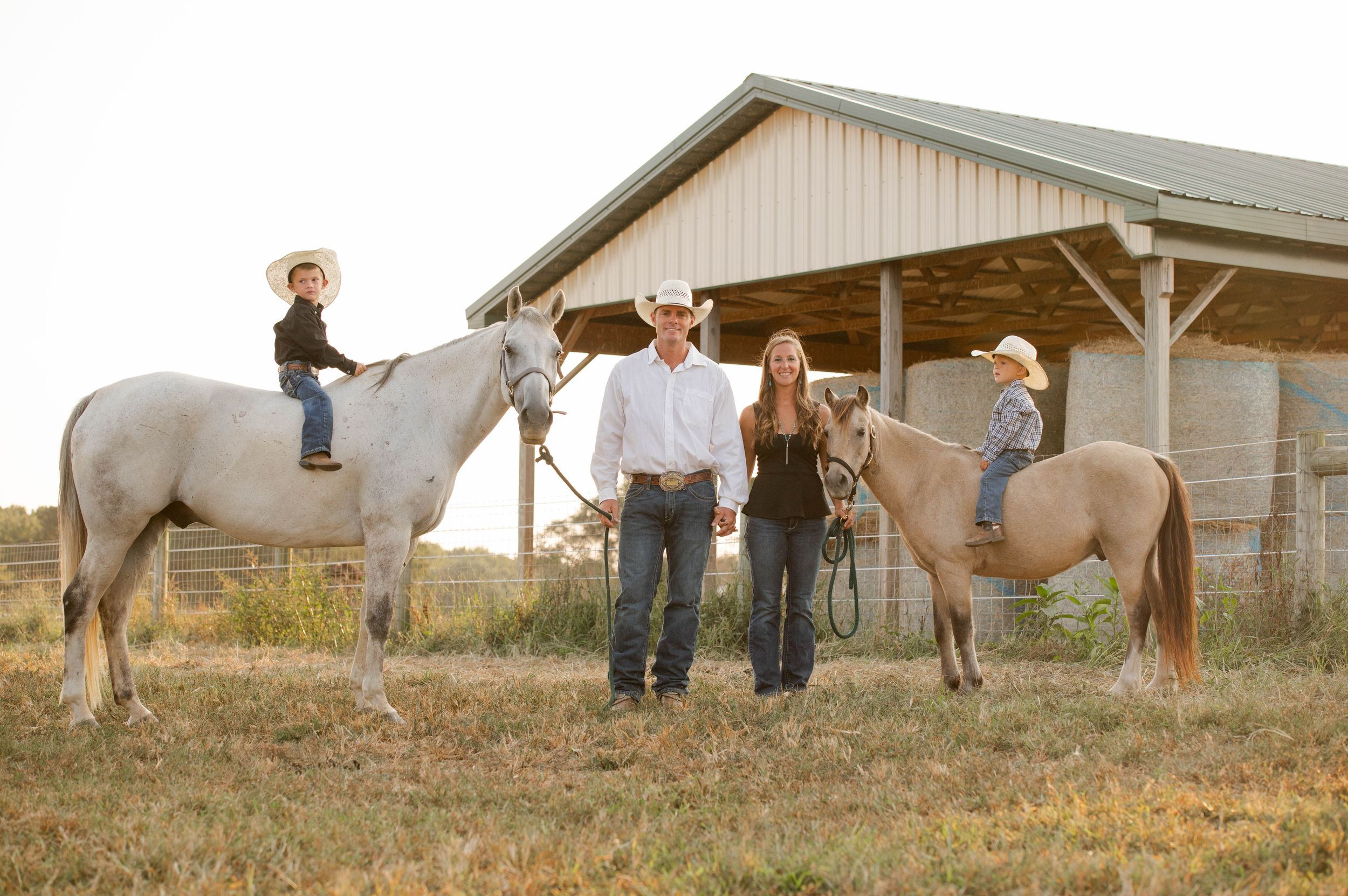 Family gathering with horses outside rustic barn at golden hour.