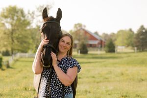 Person in polka dot dress embracing dark horse in green pasture with red barn in background.
