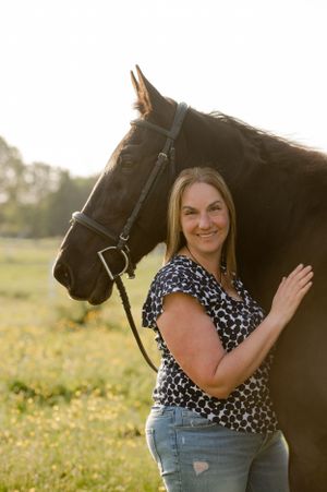 Woman in floral print top poses with black horse in sunny field.