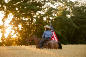 Patriotic horseback rider carrying American flag through golden field at sunset.