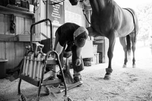 Black and white photo of farrier working on horseshoe in stable setting.