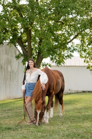 Person in white blouse and denim shorts stands with brown horse under leafy tree.