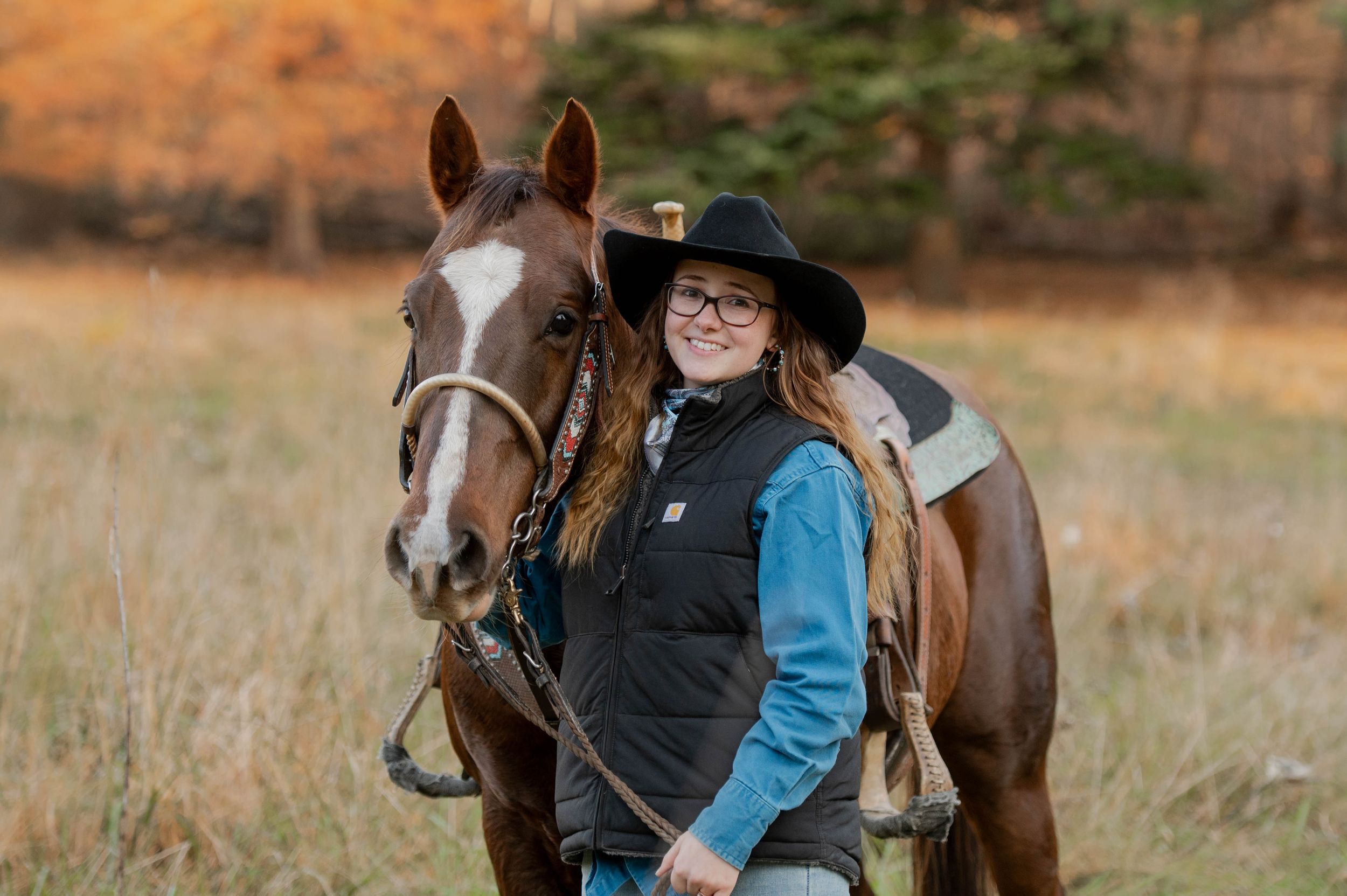 A rider in a black hat stands with a brown and white horse against fall foliage.