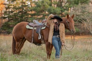 A rider in western wear stands beside a brown horse with saddle in an autumn field.