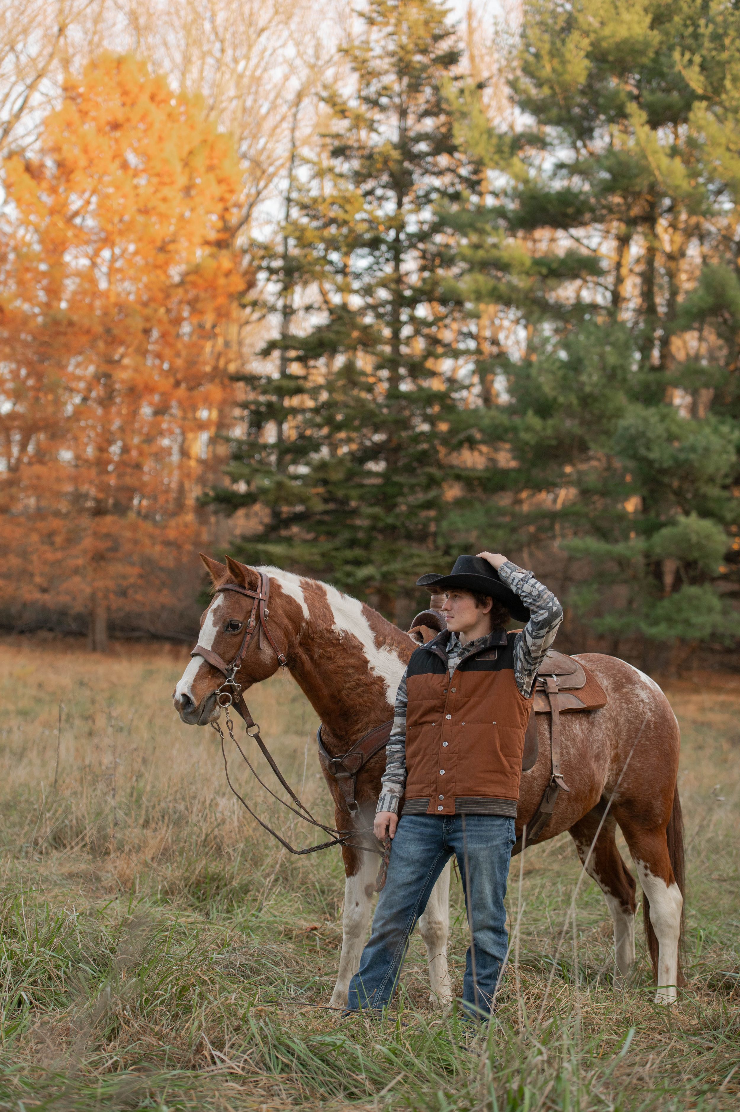 A rider stands with a paint horse against a backdrop of colorful autumn trees.