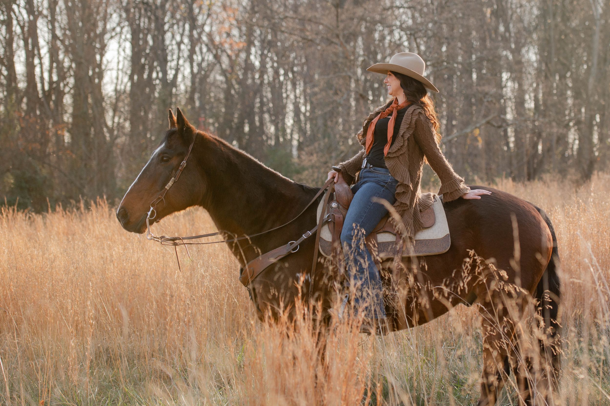 A horseback rider in western attire silhouetted against warm autumn light in tall grass.