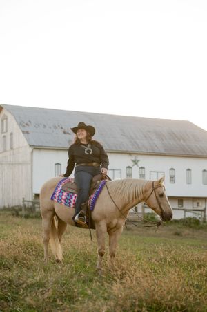 A rider on horseback poses in front of a white barn with a decorative saddle blanket.