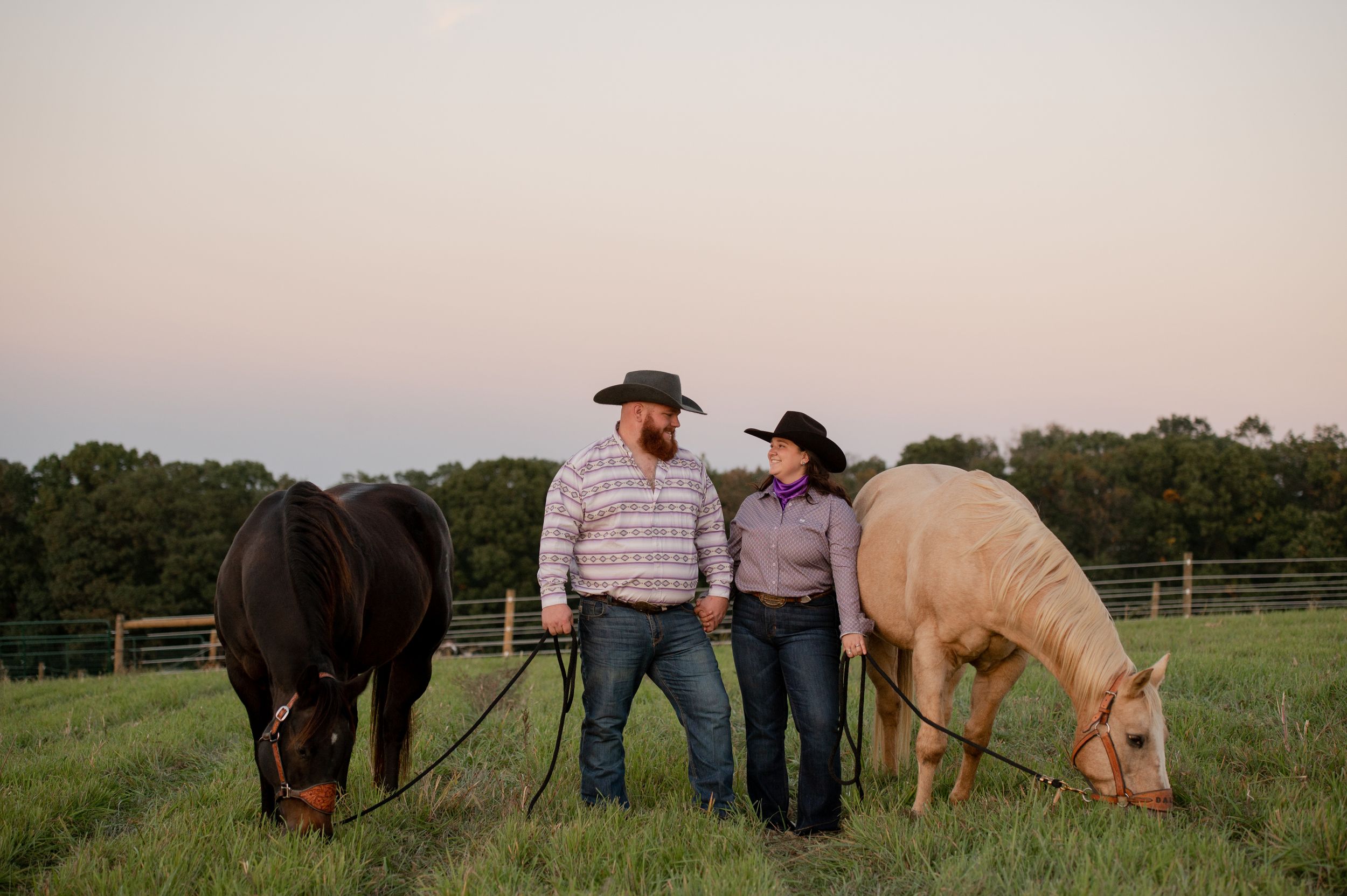Two people stand between grazing horses in a pastoral field at sunset.