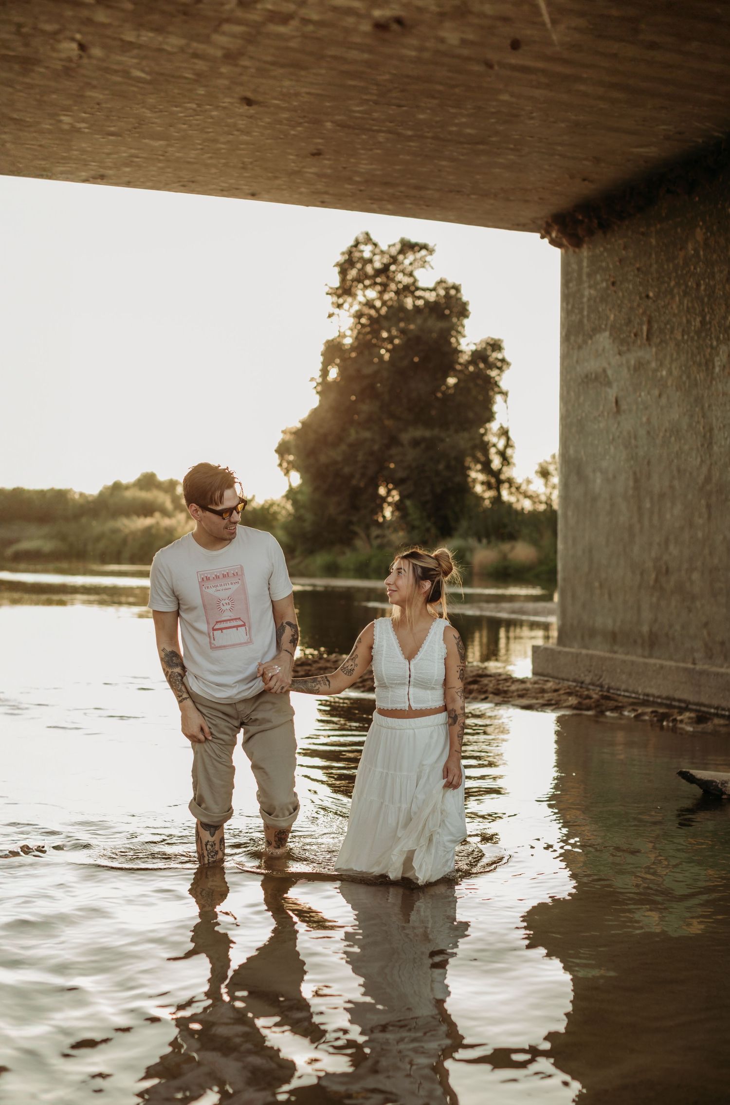 Couple wading in shallow water under bridge during sunset.