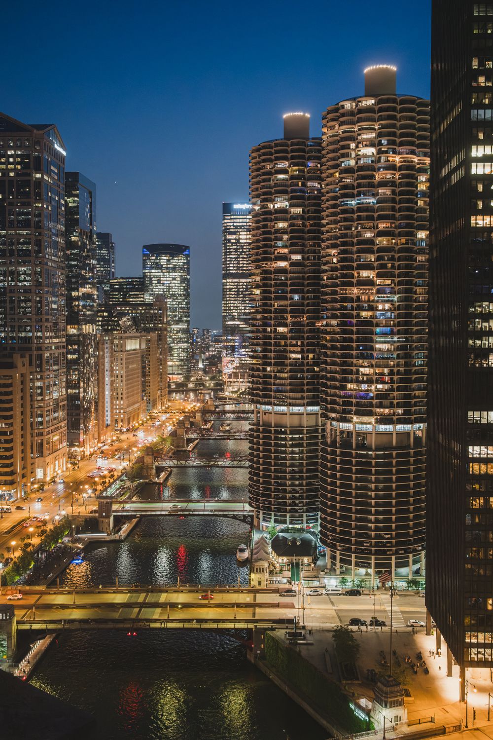 Aerial night view of Chicago River with illuminated Marina City towers and city skyline glowing under a deep blue dusk sky.