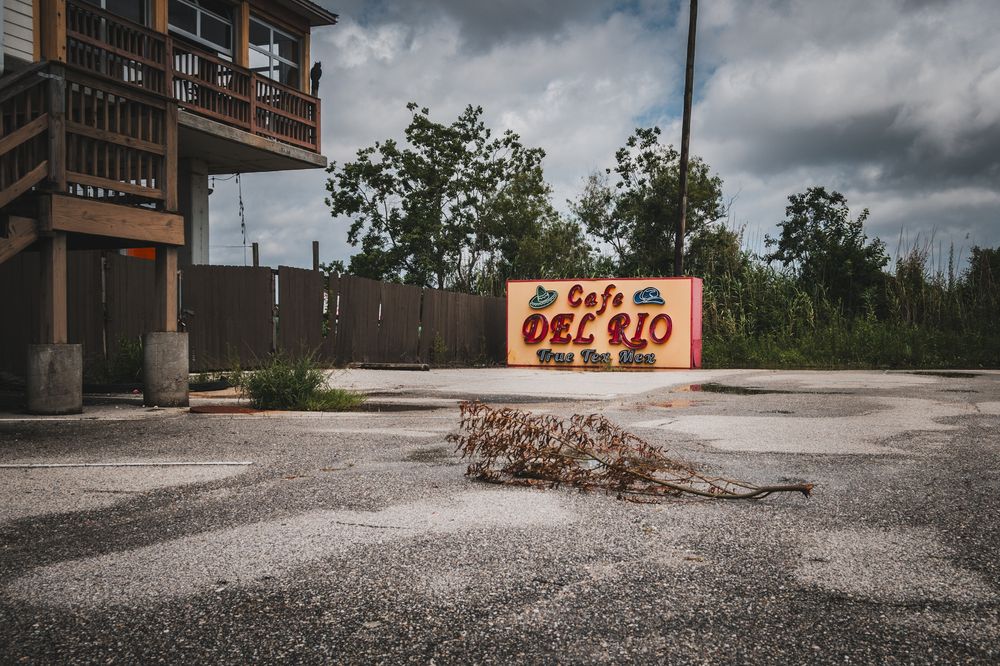 Abandoned Cafe Del Rio sign in a desolate parking lot with overgrown vegetation and a weathered building in the background.