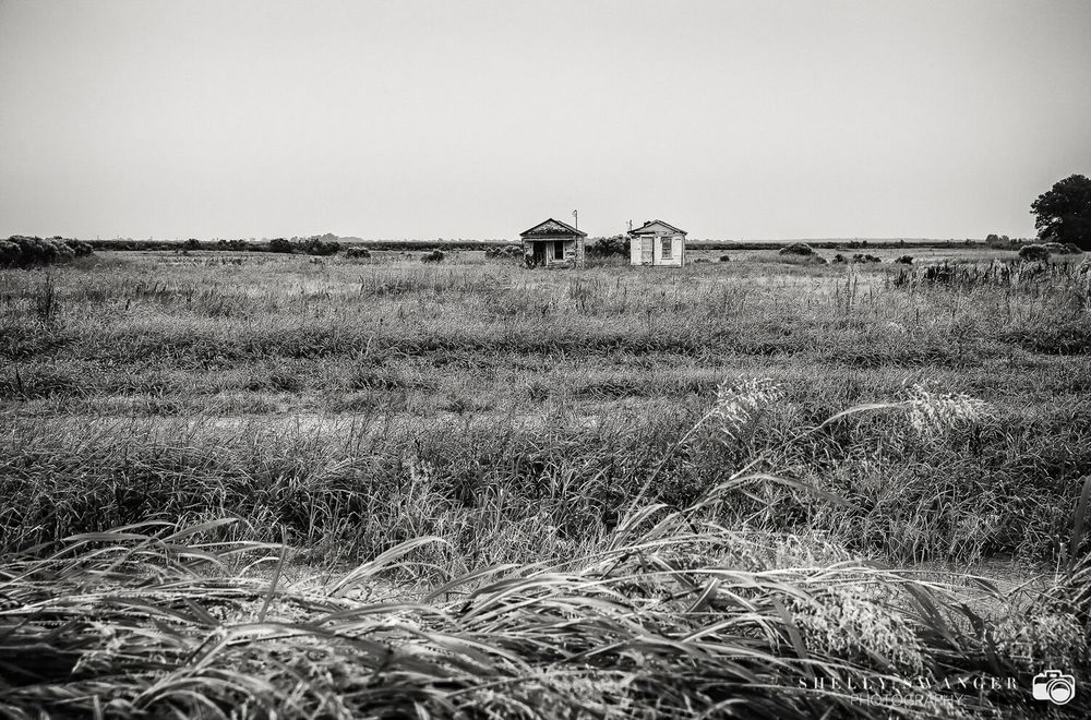 Black and white photo of abandoned rural buildings surrounded by vast open grasslands under a clear sky.